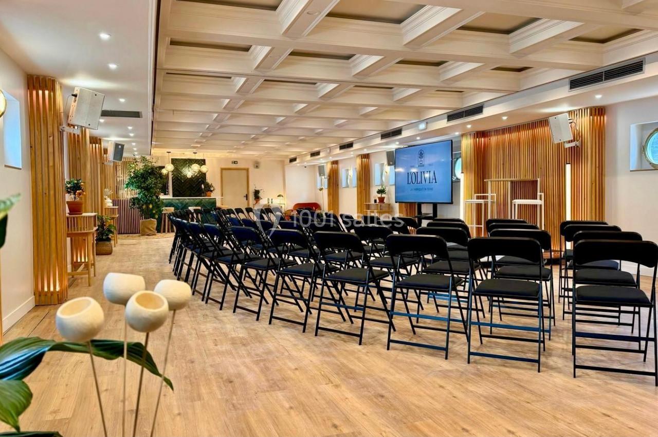 Salle de conférence lumineuse avec des rangées de chaises noires, un écran et une décoration végétale.