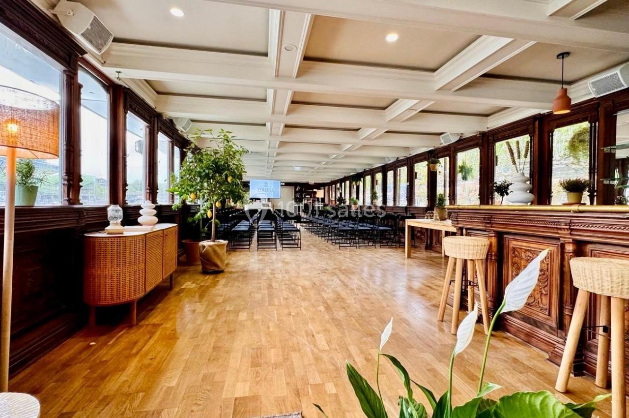 Salle lumineuse avec parquet, bar en bois, plantes décoratives et rangées de chaises disposées face à un écran.