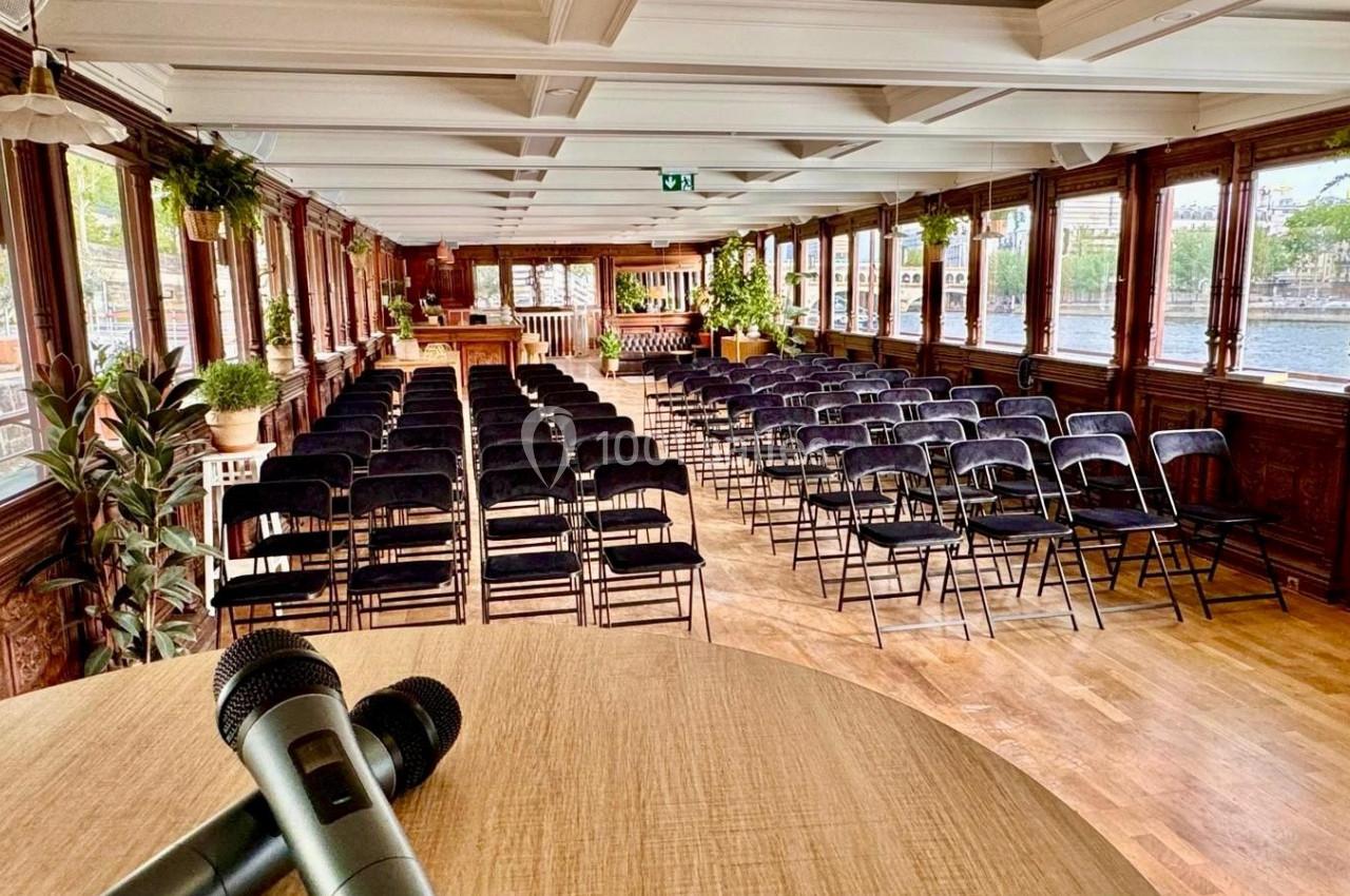 Salle de conférence lumineuse avec des rangées de chaises noires, vue depuis un pupitre avec des micros.
