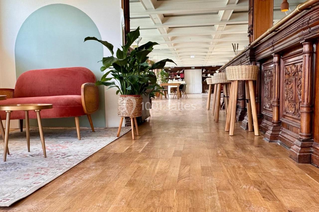 Espace intérieur lumineux avec parquet, fauteuil rouge, tables basses, plantes en pot et comptoir en bois sculpté.