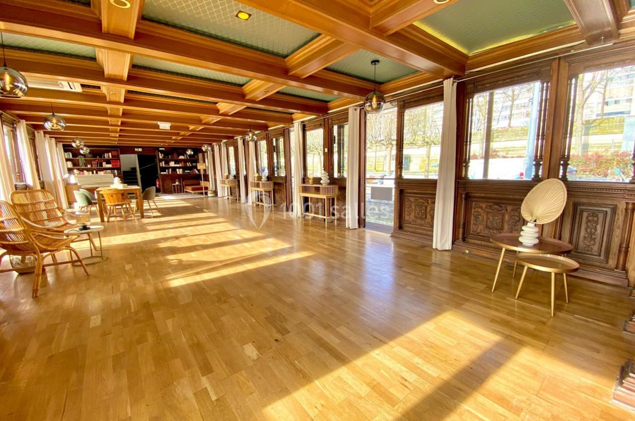 Salle lumineuse avec parquet en bois, grandes fenêtres, mobilier en bois et étagères remplies de livres au fond.
