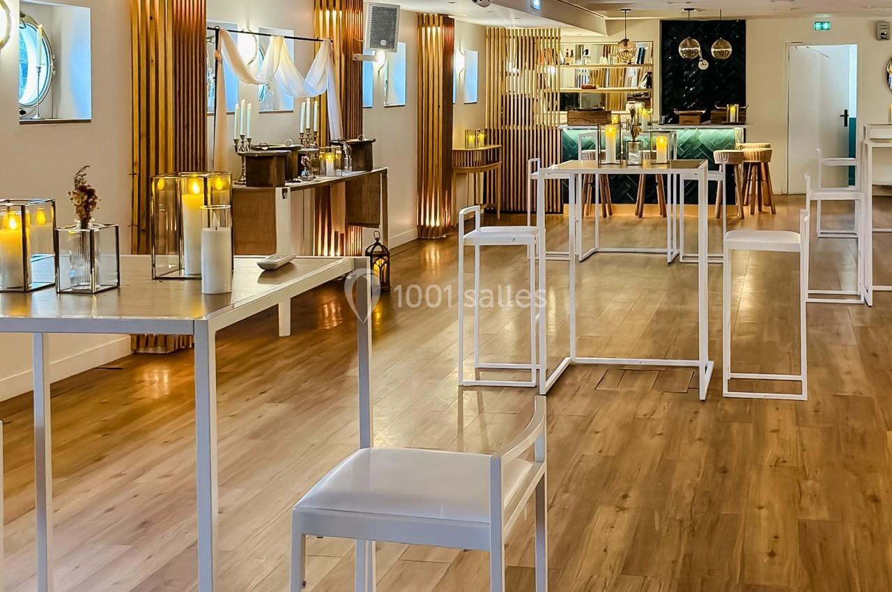 Salle lumineuse avec parquet, tables hautes blanches, bougies décoratives et un bar en arrière-plan.