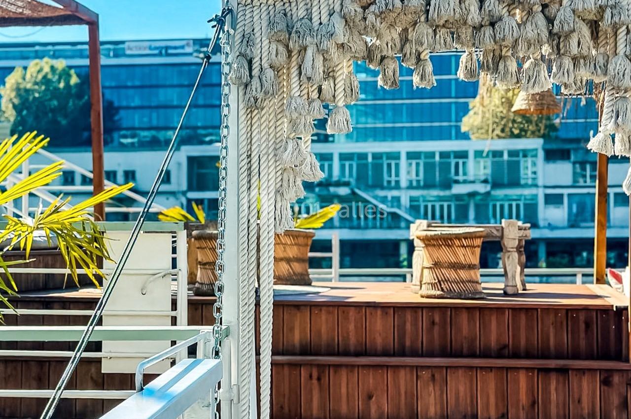 Terrasse en bois avec une arche décorative en corde, entourée de plantes et surplombant des bâtiments modernes.