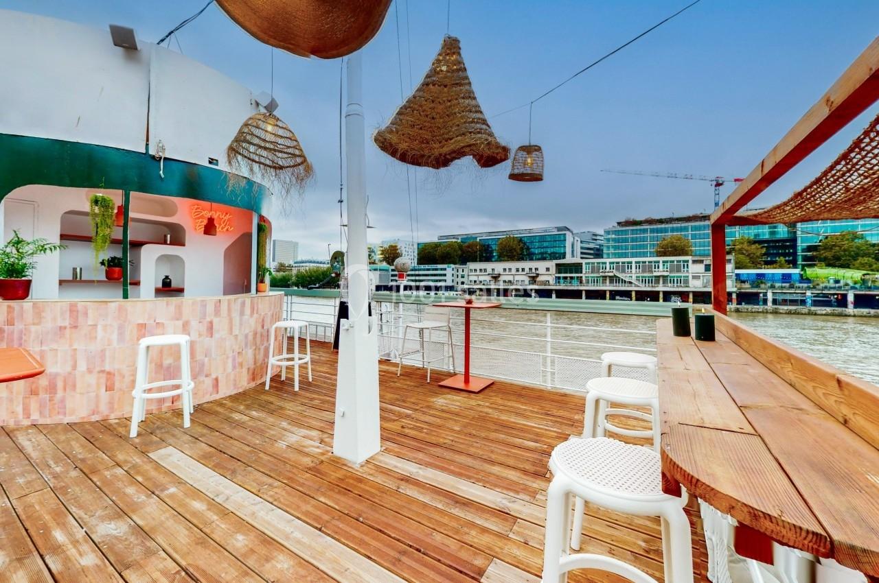 Terrasse en bois avec bar, tabourets blancs, suspensions en osier et vue sur un fleuve bordé de bâtiments modernes.