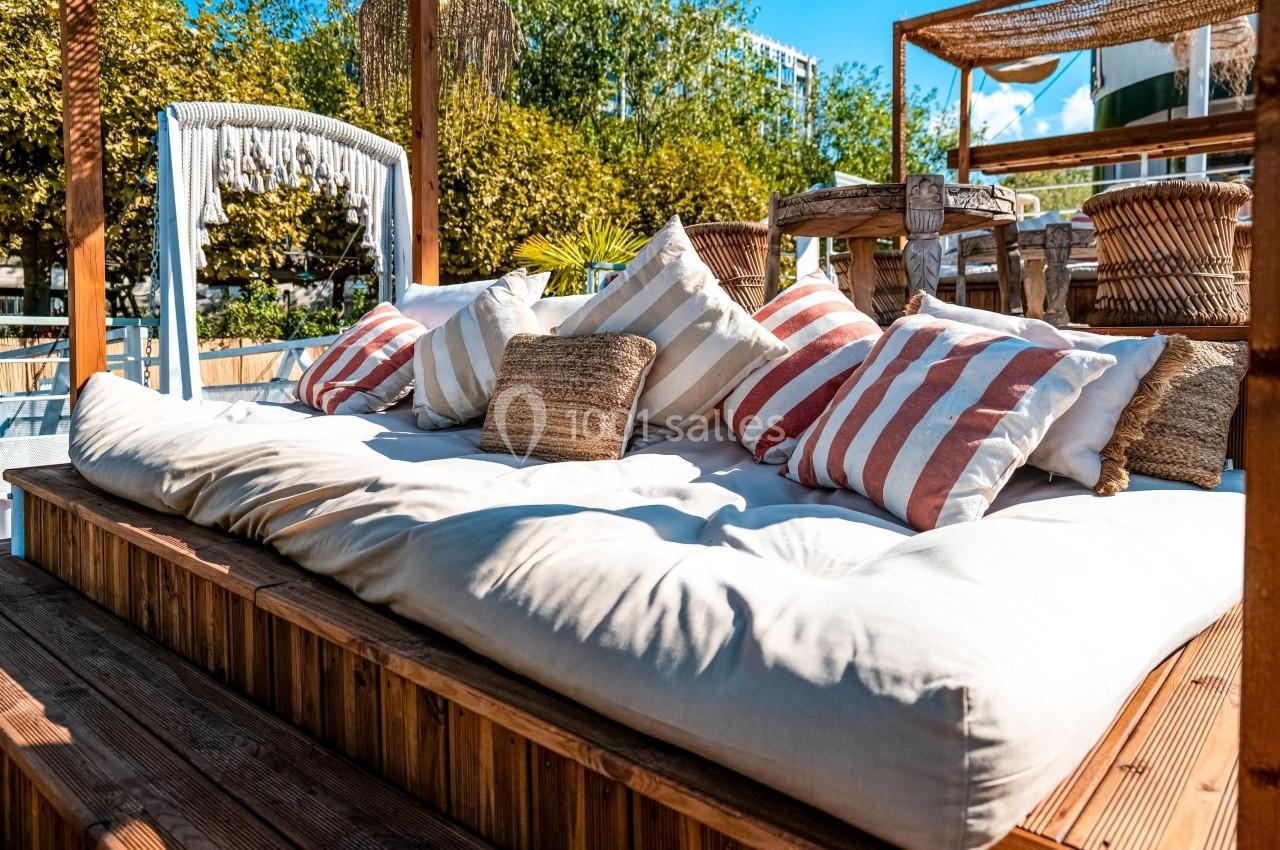 Lit de repos extérieur avec coussins rayés et un cadre en bois, entouré de végétation et mobilier en osier.