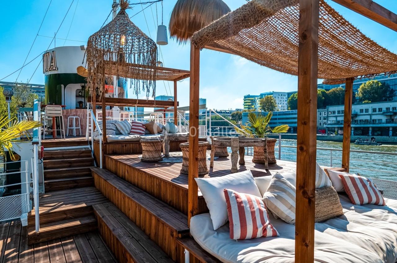 Terrasse en bois aménagée avec coussins, fauteuils et parasols, donnant sur un fleuve et des bâtiments urbains.