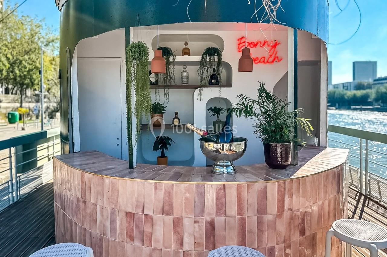 Bar extérieur avec comptoir en carreaux roses, plantes décoratives, bouteilles exposées et vue sur un fleuve en arrière-plan.