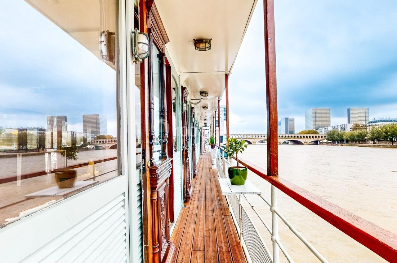 Vue d'une péniche avec un ponton en bois, longeant une rivière et des bâtiments modernes en arrière-plan.