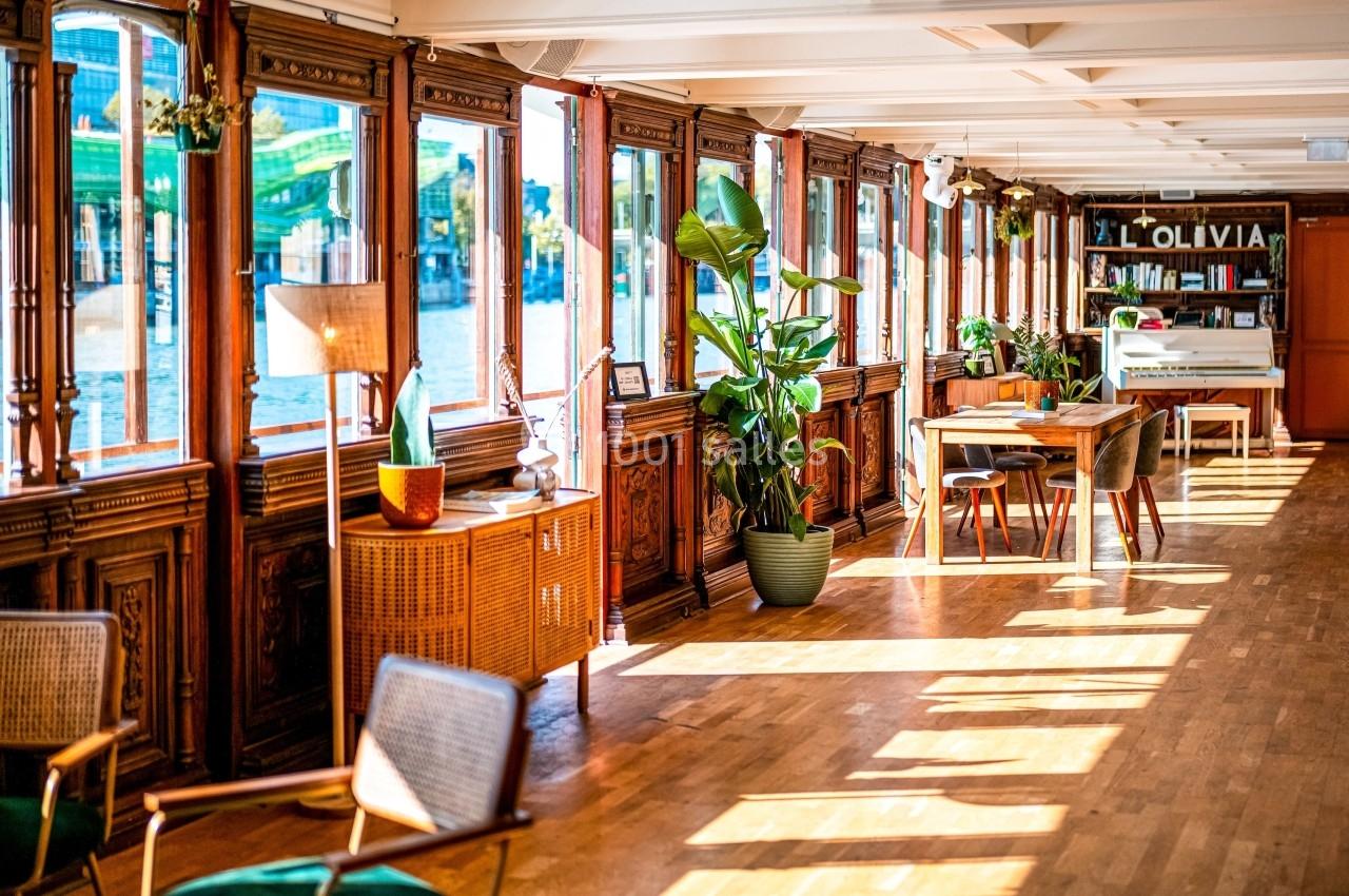 Salle lumineuse avec parquet, grandes fenêtres, mobilier en bois et plantes, offrant une vue sur l'extérieur.