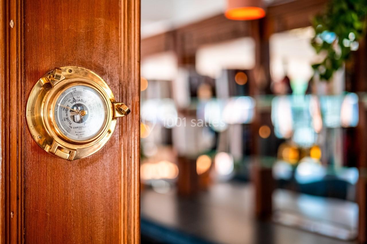 Baromètre en laiton fixé sur une paroi en bois, avec un arrière-plan flou d'intérieur lumineux et décoré.