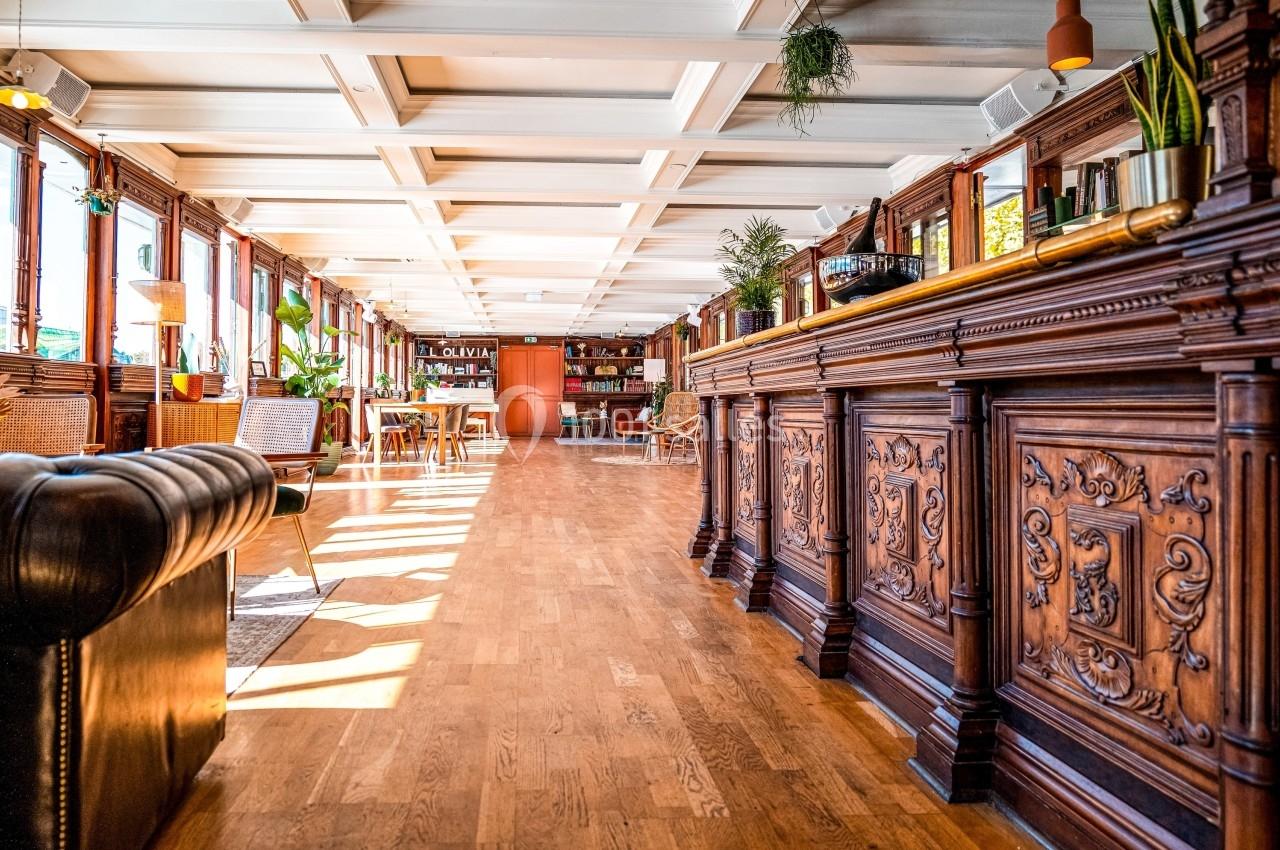 Intérieur lumineux avec parquet, comptoir en bois sculpté, fauteuils et plantes dans un espace chaleureux.