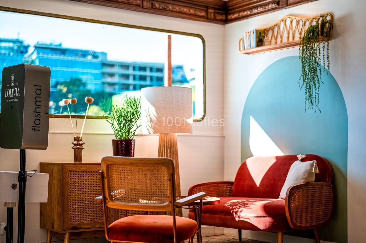 Espace lumineux avec fauteuils en rotin, lampe, plantes et fenêtre donnant sur des bâtiments modernes.