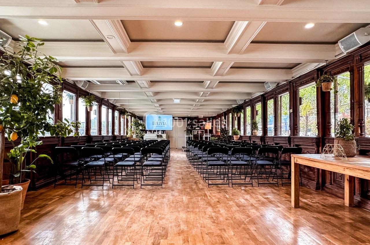 Salle lumineuse avec parquet en bois, rangées de chaises noires, plantes en pot et grandes fenêtres donnant sur l'extérieur.
