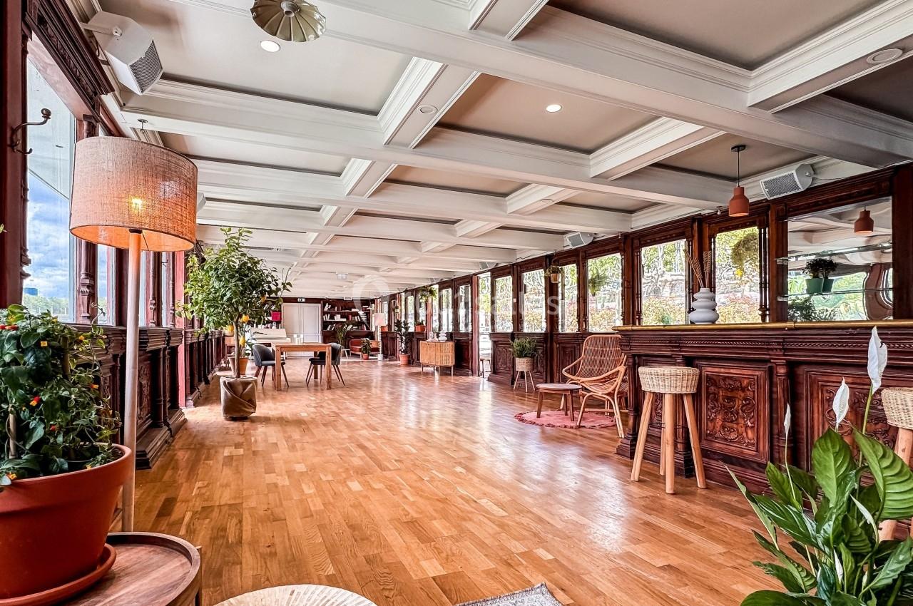 Intérieur lumineux avec parquet en bois, grandes fenêtres, plantes et mobilier en bois dans un espace ouvert.