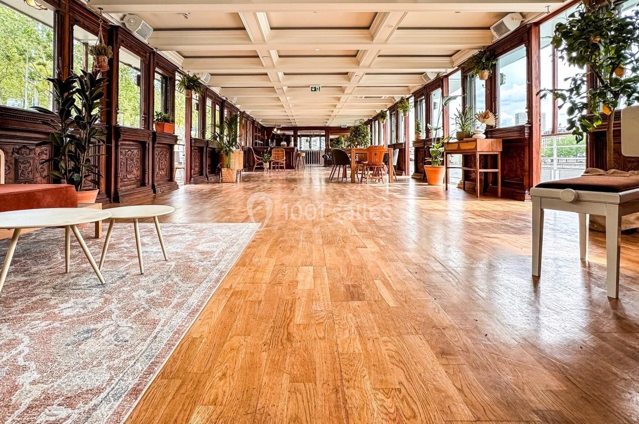 Salle lumineuse avec parquet en bois, grandes fenêtres, plantes et mobilier varié, offrant une vue sur l'extérieur.