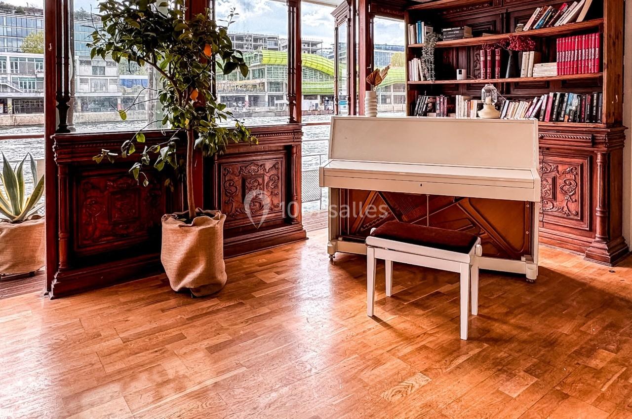 Piano blanc avec tabouret dans une pièce lumineuse, entouré de plantes et d'étagères remplies de livres.