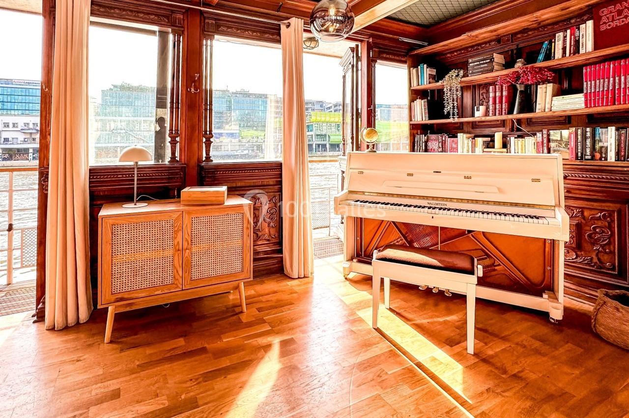 Intérieur lumineux avec piano blanc, bibliothèque en bois, meuble vintage et vue sur l'eau à travers de grandes fenêtres.