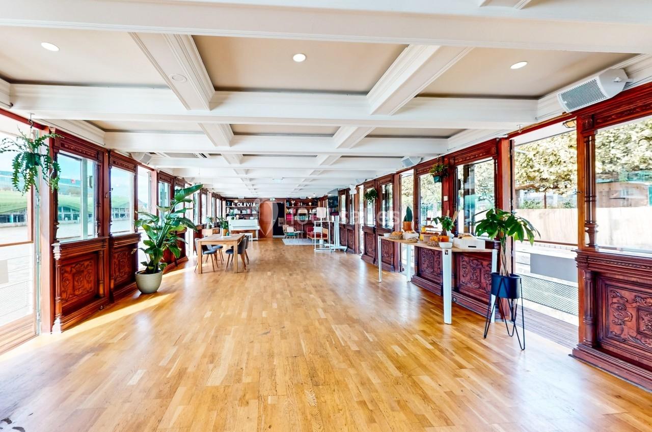 Salle lumineuse avec parquet en bois, grandes fenêtres, plantes et étagères au fond.