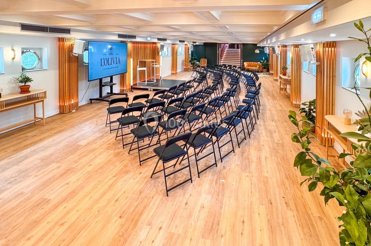 Salle lumineuse avec rangées de chaises noires, écran de présentation, parquet clair et décorations végétales.