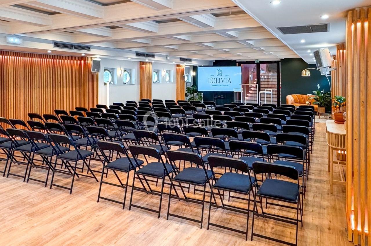 Salle de conférence lumineuse avec rangées de chaises noires alignées face à un écran de présentation.
