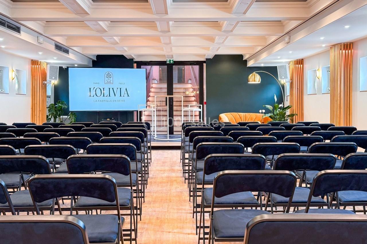 Salle de conférence lumineuse avec rangées de chaises noires, écran de projection et décor moderne.