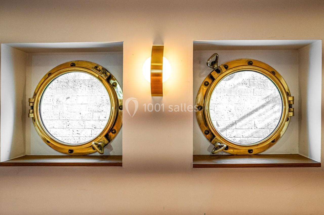 Deux hublots ronds en laiton encastrés dans un mur clair, éclairés par une applique centrale.