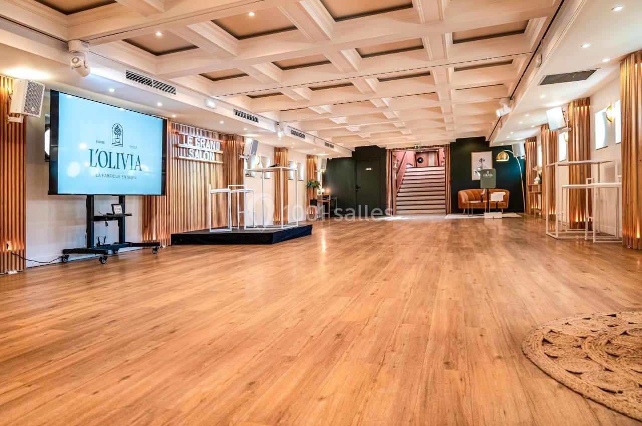 Salle lumineuse avec parquet en bois, écran de présentation, estrade et escaliers menant à un étage supérieur.