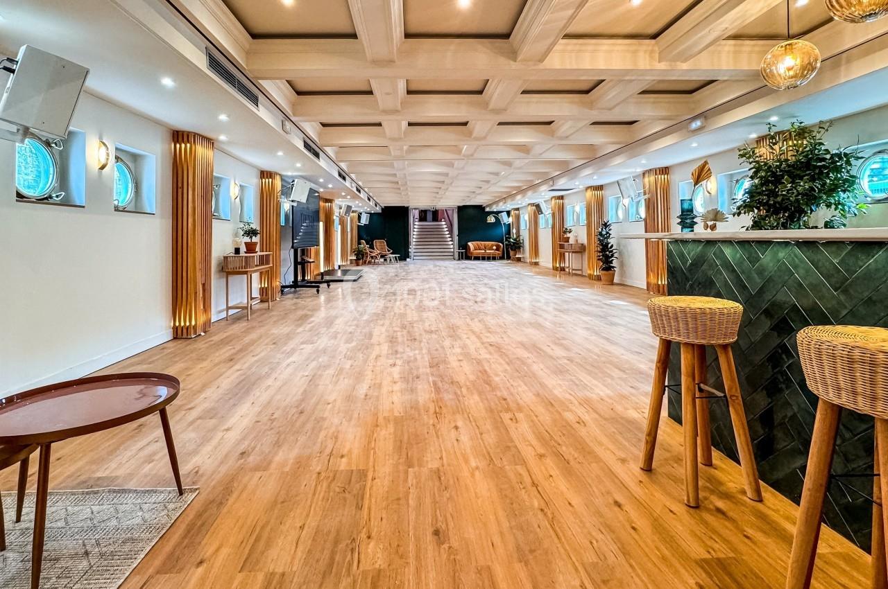 Salle spacieuse avec parquet en bois, bar à droite, luminaires suspendus et décorations végétales.