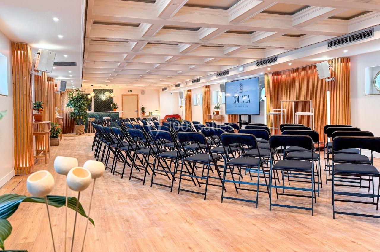 Salle lumineuse avec chaises noires alignées, écran de présentation, parquet clair et décoration végétale.