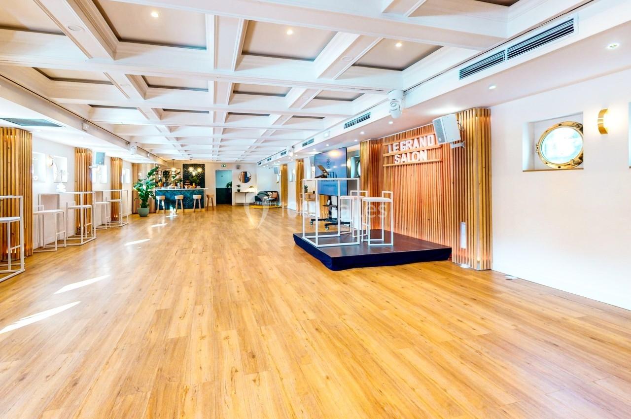 Salle lumineuse avec parquet en bois, estrade centrale, murs blancs et hublots, décorée de plantes et éclairages modernes.