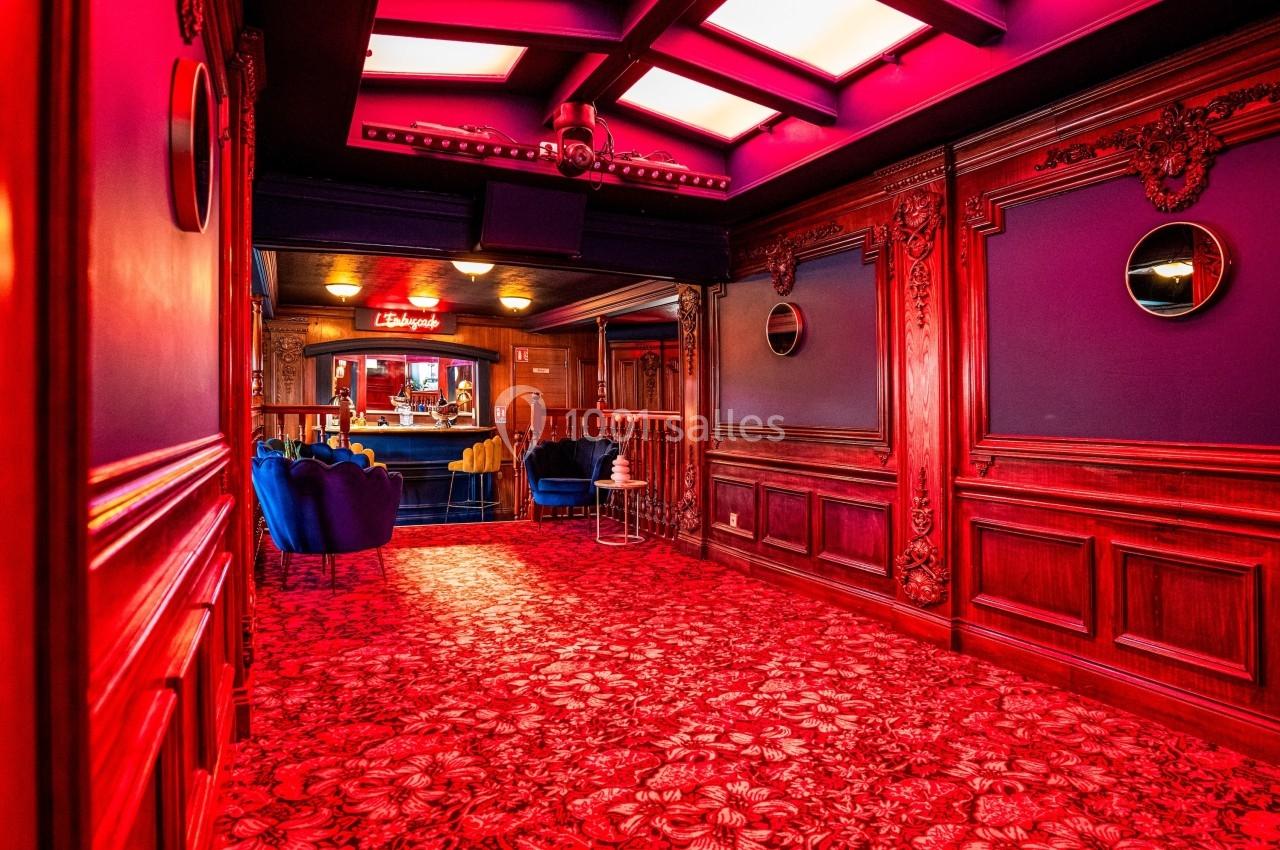 Couloir richement décoré avec tapis rouge à motifs, boiseries sculptées, et vue sur un espace lounge éclairé.