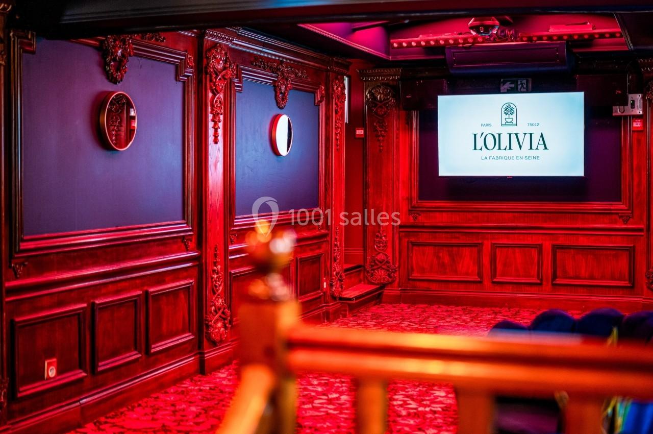 Salle décorée de boiseries rouges avec écran affichant ’L'Olivia’, sièges en bas et miroirs muraux.