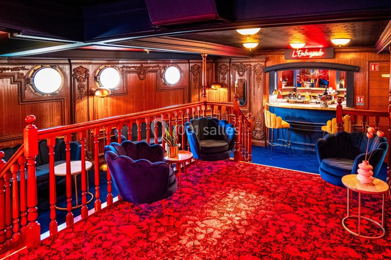 Salon chaleureux avec fauteuils bleus, tapis rouge à motifs, boiseries sculptées et bar rétro éclairé au fond.