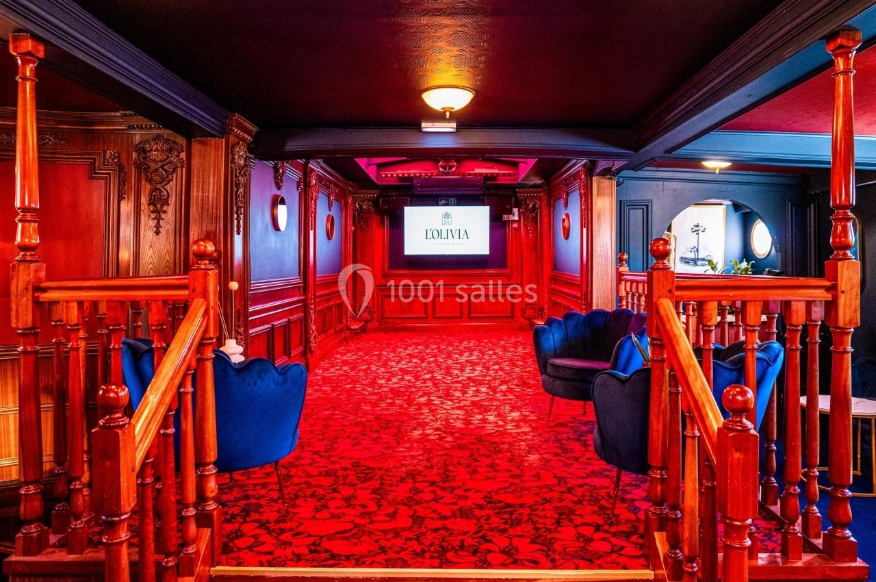 Salle élégante avec tapis rouge, fauteuils bleus, boiseries sculptées et écran affichant un logo au fond.