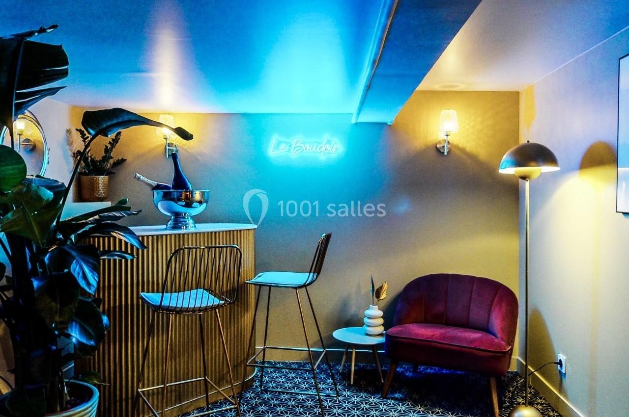 Coin salon avec fauteuil rose, bar en bois, plantes et éclairage néon bleu dans une ambiance tamisée.