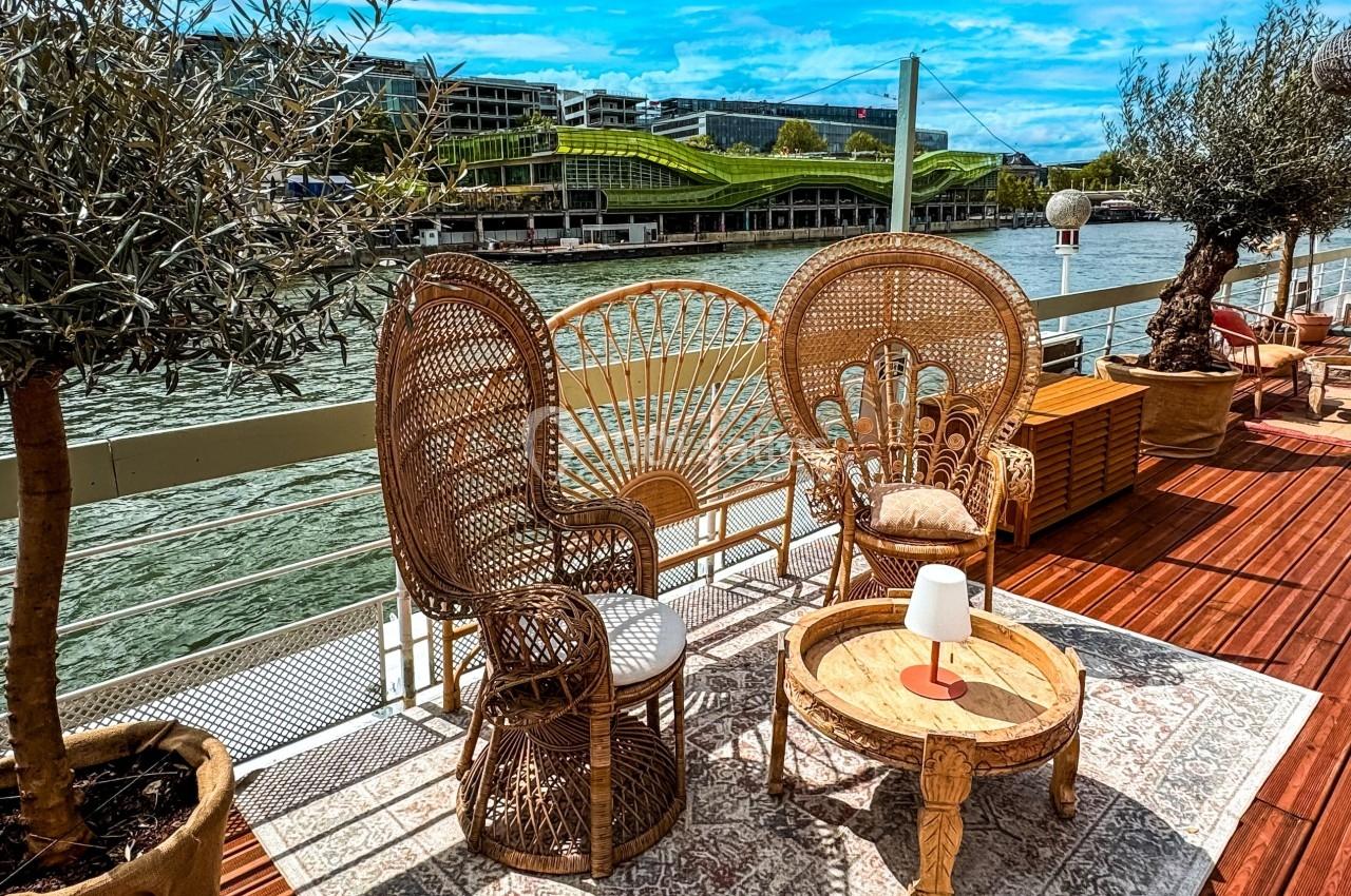 Mobilier en rotin disposé sur une terrasse en bois au bord d'une rivière, avec vue sur un bâtiment moderne et un pont.