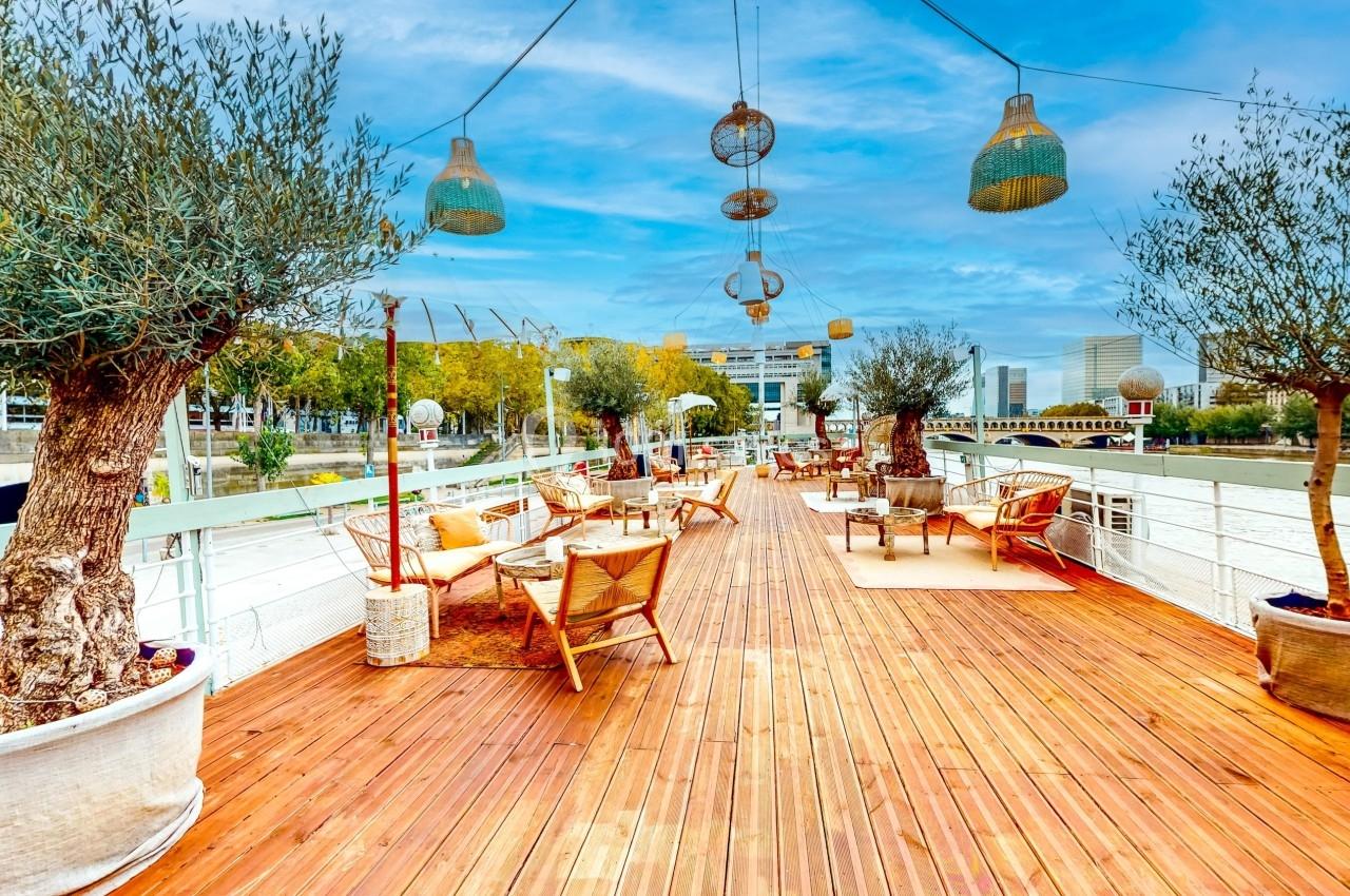 Terrasse en bois avec chaises et tables, entourée d'oliviers en pots, surplombant une rivière par temps clair.