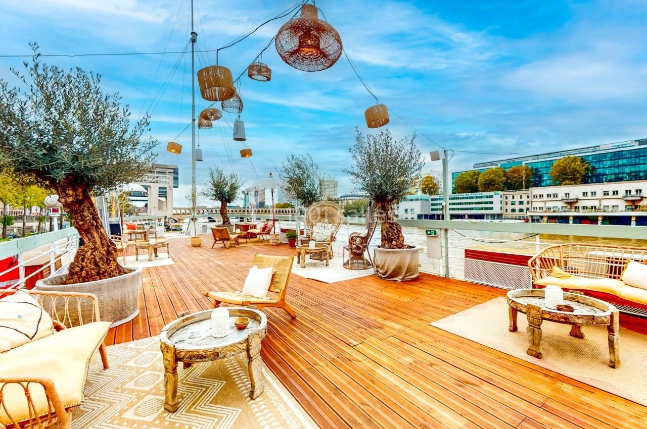 Terrasse en bois aménagée avec des fauteuils, tables et oliviers, surplombant un paysage urbain près d'un cours d'eau.