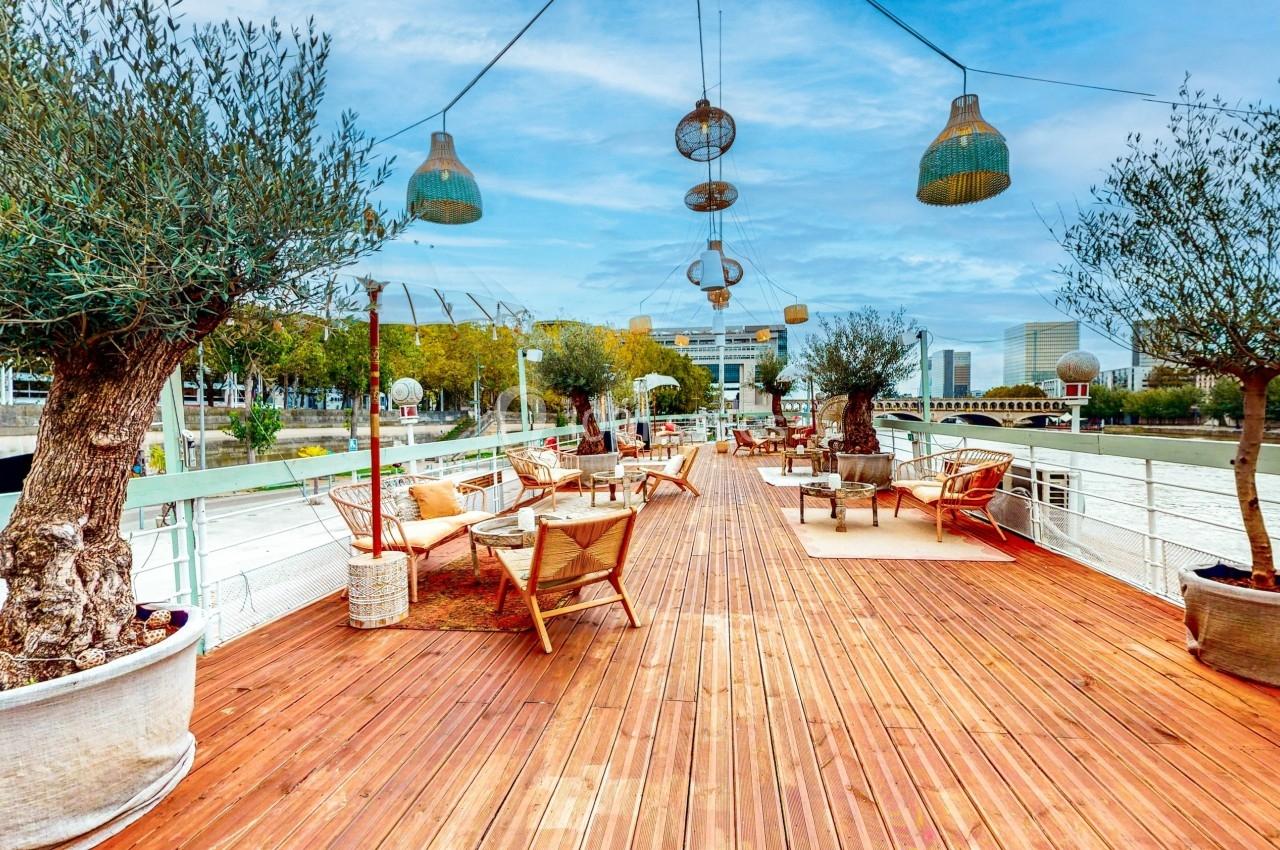 Terrasse en bois aménagée avec des fauteuils, des tables et des oliviers en pots, surplombant un cours d'eau.