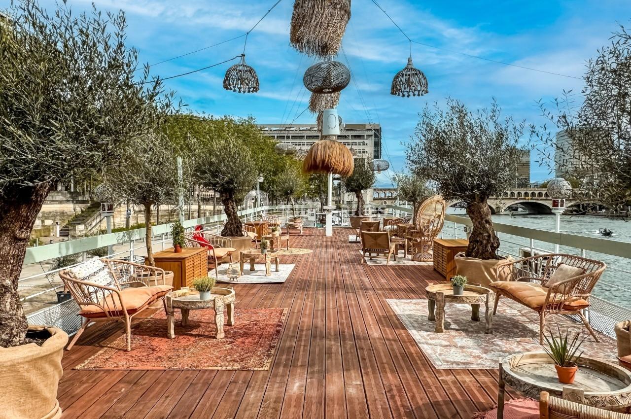 Terrasse en bois aménagée avec des fauteuils, tapis et plantes, surplombant une rivière sous un ciel bleu.
