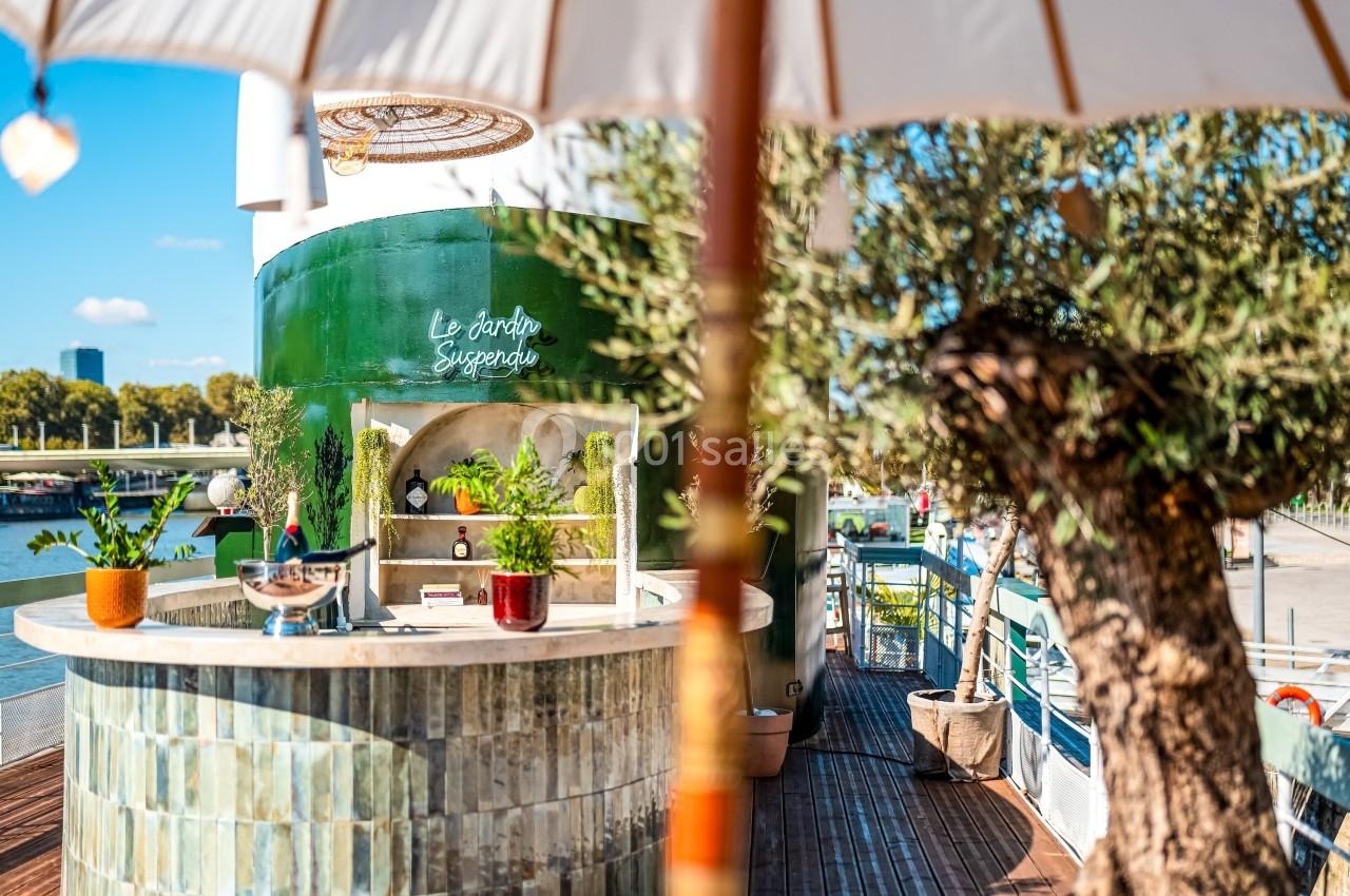 Bar extérieur en céramique verte entouré de plantes, situé sur une terrasse ensoleillée près d’un fleuve.