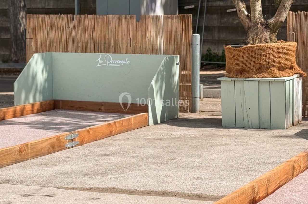 Terrain de pétanque en extérieur avec bordures en bois, sol en béton et un olivier en pot en arrière-plan.