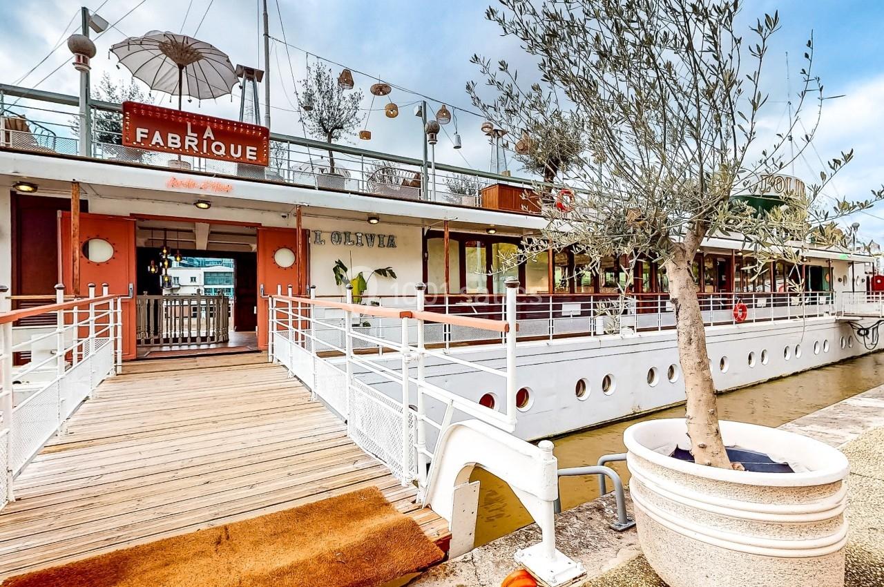 Entrée d'un restaurant aménagé sur un bateau, avec une passerelle en bois et des plantes en pot.