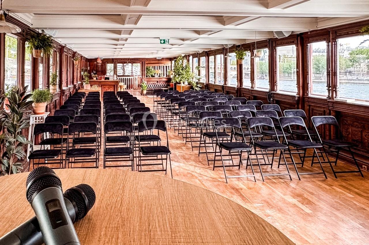 Salle de conférence lumineuse sur une péniche, avec des rangées de chaises noires et des micros sur une table au premier…