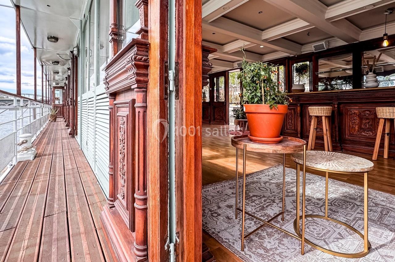 Vue d'un intérieur lumineux avec mobilier en bois sculpté, plante en pot sur une table et terrasse donnant sur l'eau.