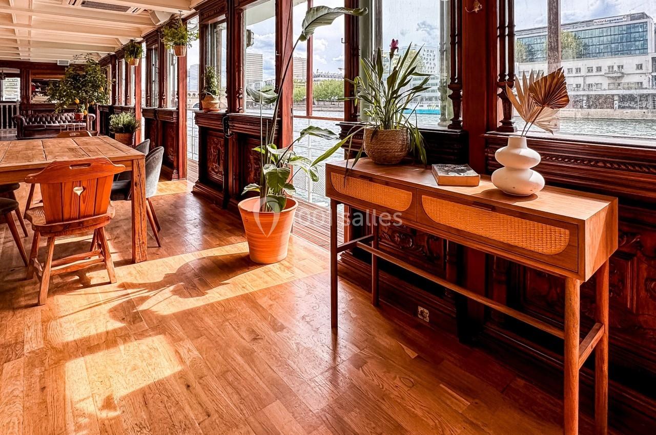 Intérieur lumineux avec parquet, meubles en bois, plantes et vue sur l'eau à travers de grandes fenêtres.