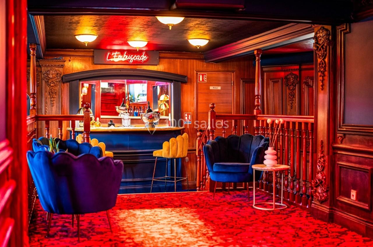 Salon chaleureux avec fauteuils colorés, bar en arrière-plan et enseigne lumineuse rouge indiquant ’L'Ambuscade’.