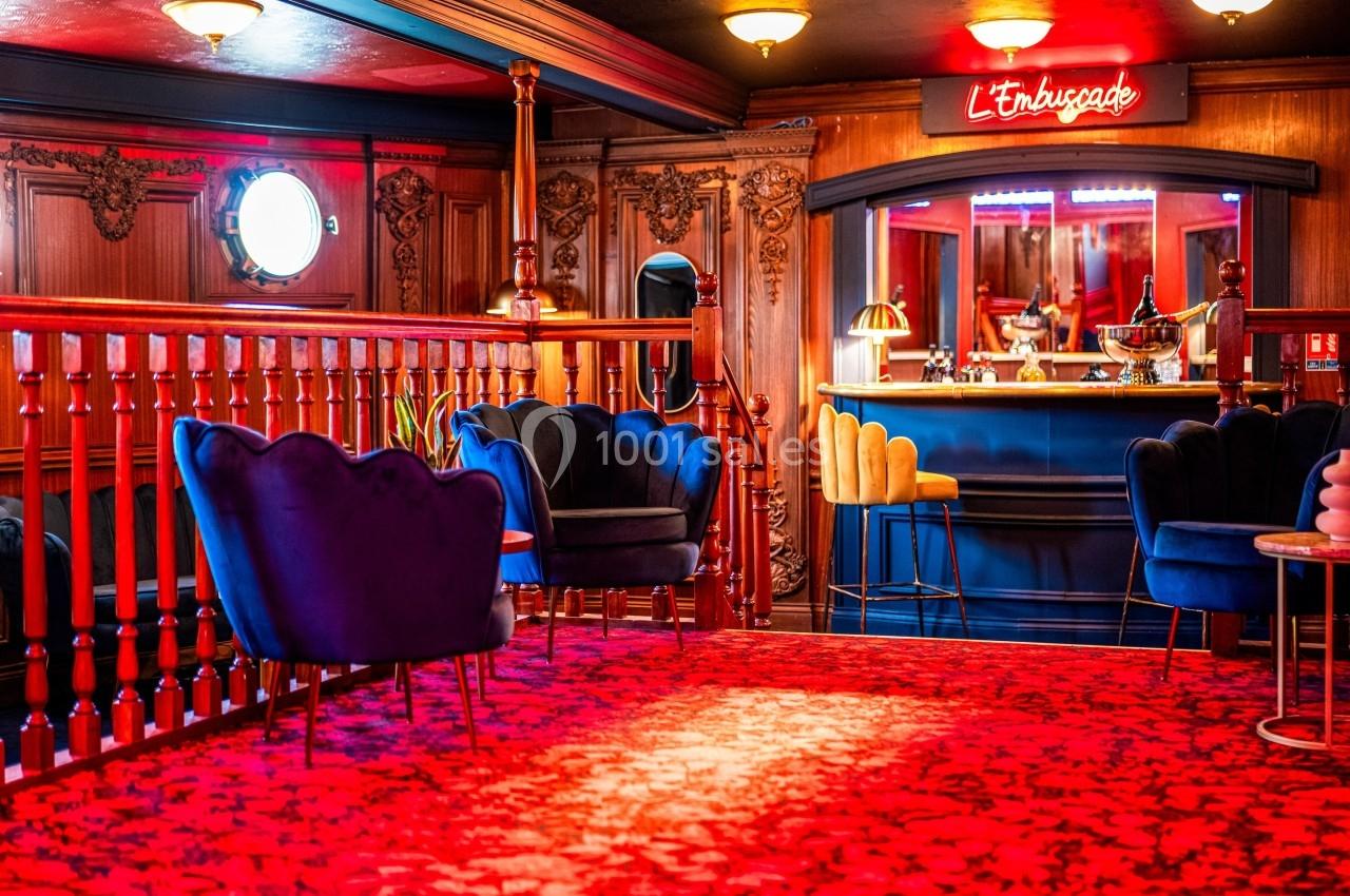 Salon avec fauteuils colorés, tapis rouge à motifs, boiseries sculptées et bar éclairé par une enseigne néon.