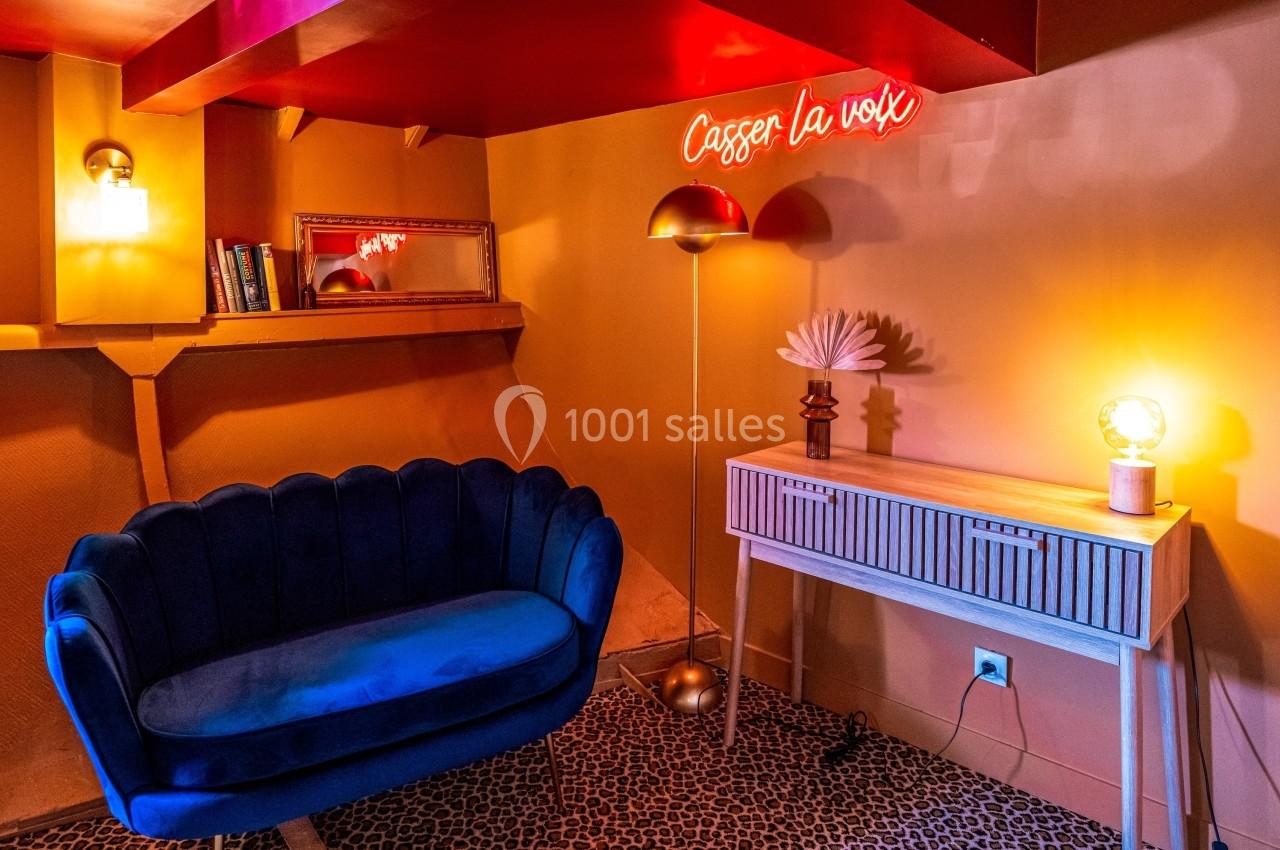 Salon cosy avec canapé bleu, lampes design, console blanche et néon rouge affichant ’Casser la voix’.