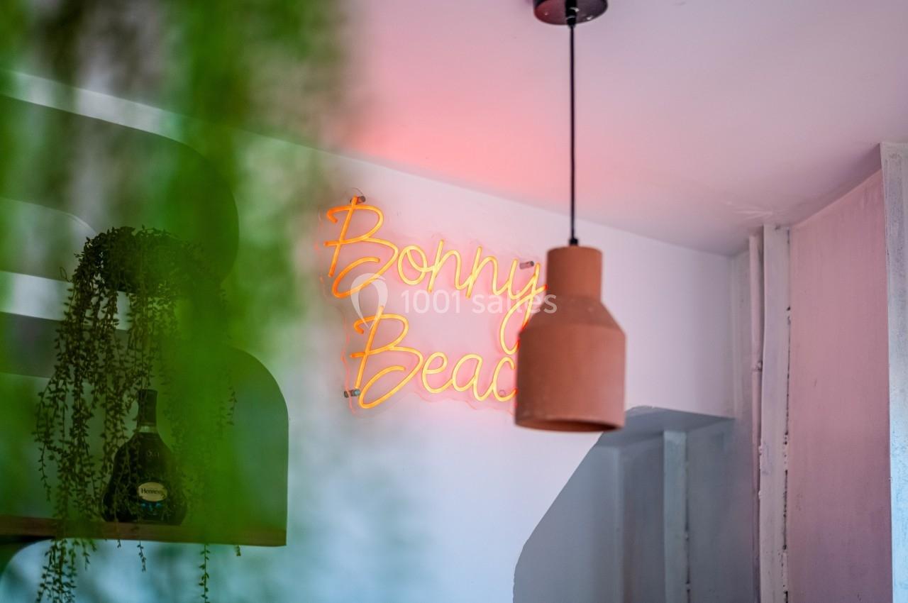 Enseigne néon ’Bonny Beach’ accrochée à un mur blanc, avec une suspension et des plantes en premier plan.
